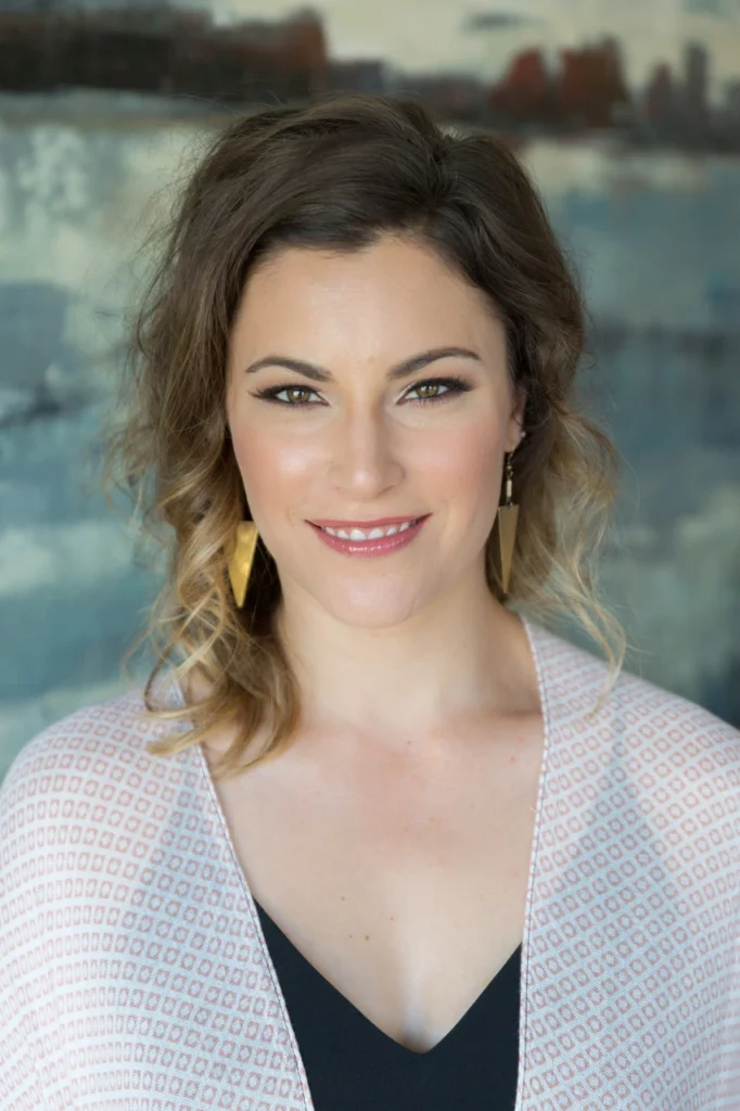 A woman with shoulder-length wavy hair, wearing a light patterned top and gold earrings, smiles at the camera against a blurred background.