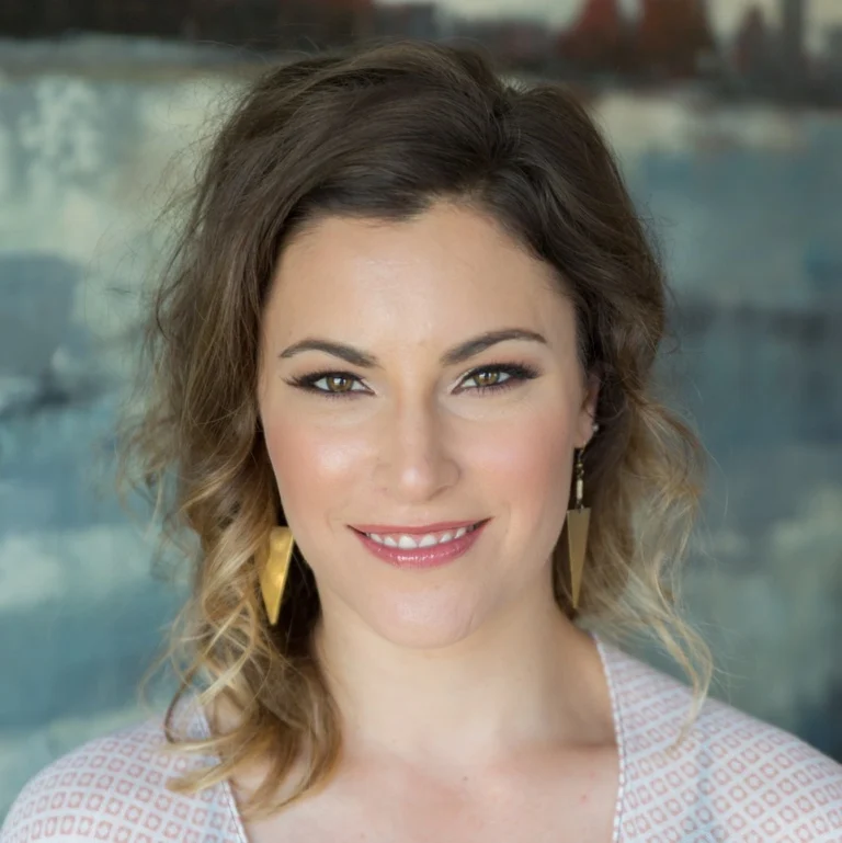 A woman is posing for a photo in front of a blurred, abstract background. She has shoulder-length wavy hair, wears earrings, and a light-colored top. She is looking at the camera with a slight smile.