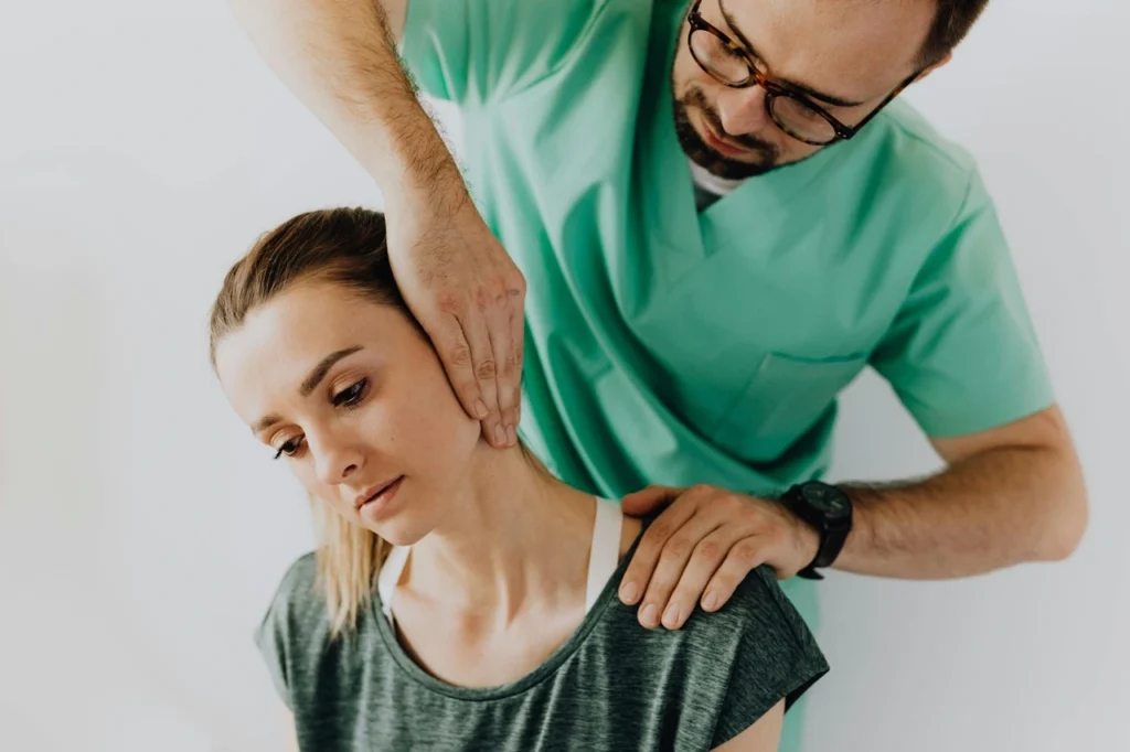 A person in a green medical uniform assists a seated woman by gently stretching her neck to the side.