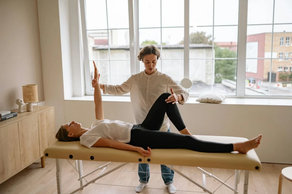 Person lying on a massage table with another person assisting in an arm and leg stretch, in a bright room with large windows.